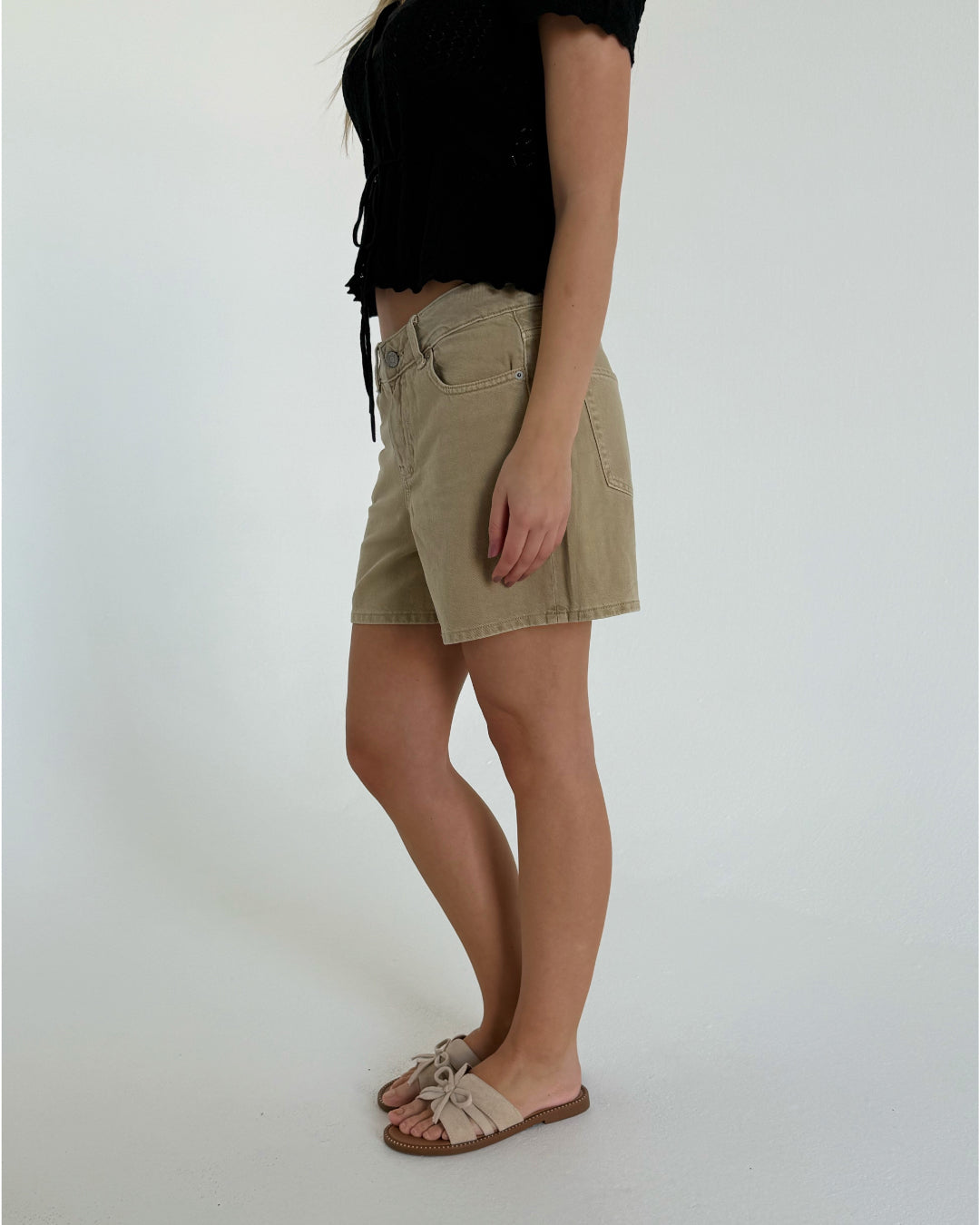 En person iført en sort bluse og beige shorts står i profil, iført lyse sandaler med sløjfer. Baggrunden er en ensfarvet, lys væg.