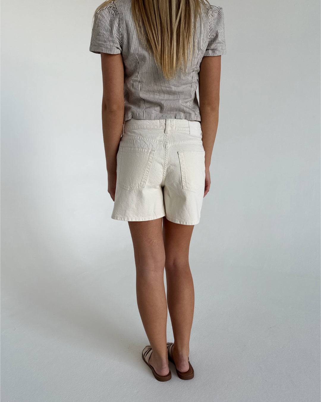 En person iført stribet top og hvide shorts står med ryggen til, iført sandaler, i et lyst rum.