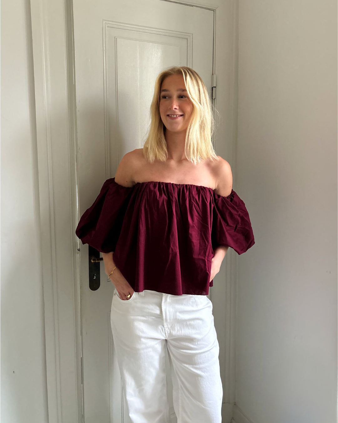En person står smilende iført en mørkerød, skulderløs top og hvide bukser, med hænderne i lommerne, foran en hvid dør i et lyst rum.