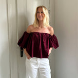 En person står smilende iført en mørkerød, skulderløs top og hvide bukser, med hænderne i lommerne, foran en hvid dør i et lyst rum.