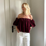 En person står i en gang iført en burgundy off-shoulder bluse og hvide bukser. Baggrunden viser en hvid dør og vægge.