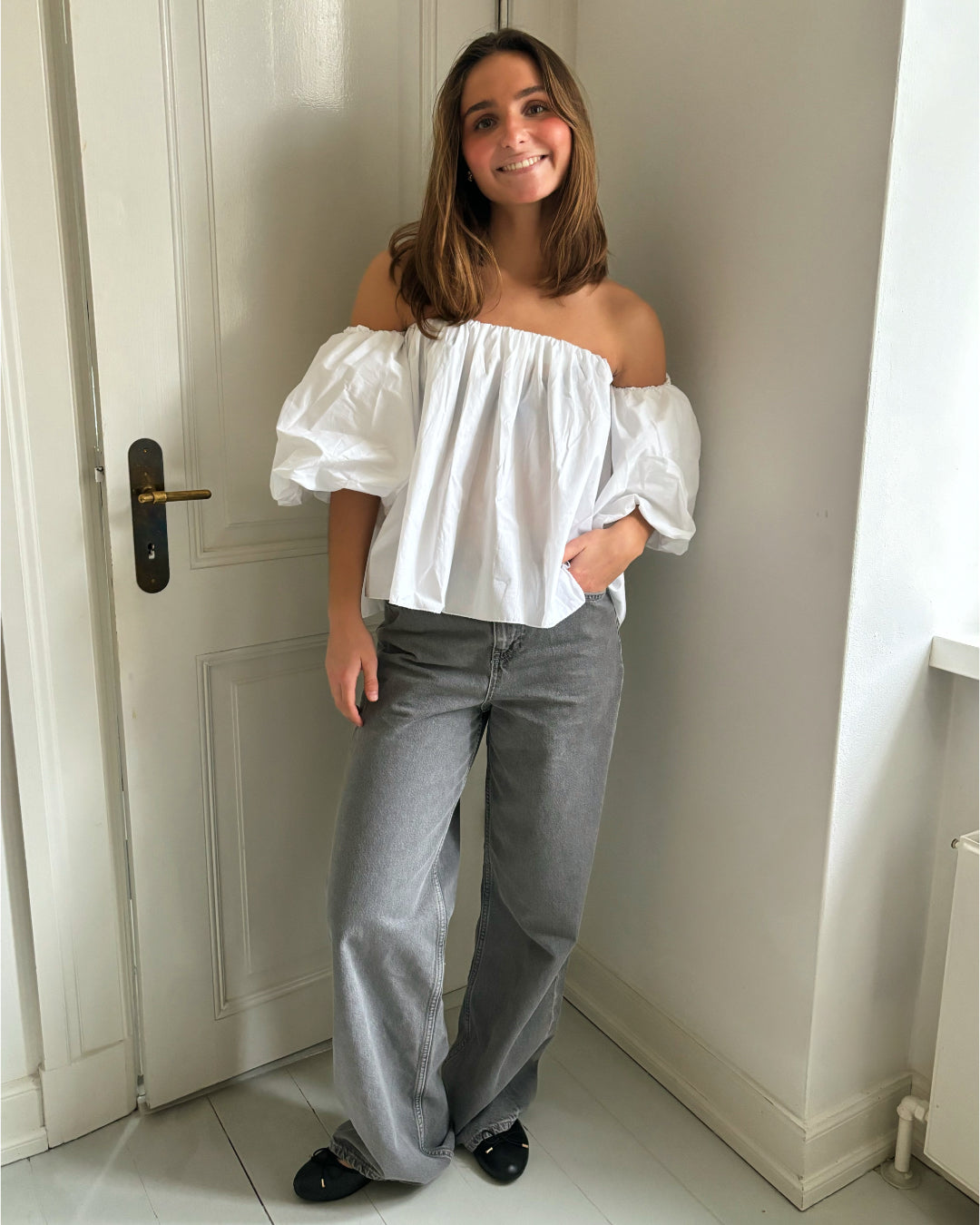 En person står afslappet i et rum med lyse vægge og en hvid dør. Personen bærer en hvid off-shoulder-bluse og grå bukser, og de smiler.