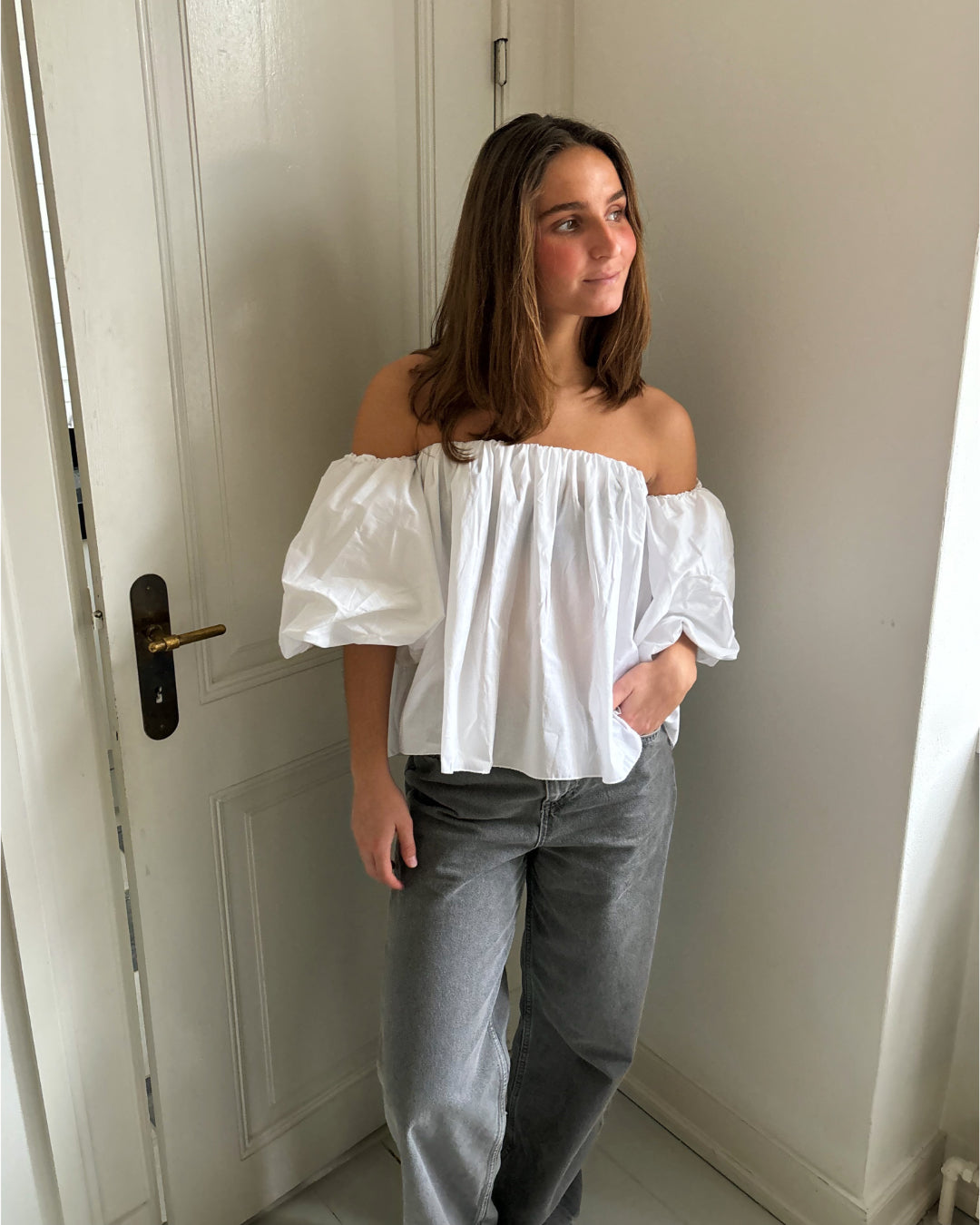 En person står ved en hvid dør iført en hvid off-shoulder bluse og grå bukser. Rummet er lyst med en enkel indretning.