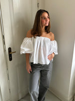 En person står ved en hvid dør iført en hvid off-shoulder bluse og grå bukser. Rummet er lyst med en enkel indretning.