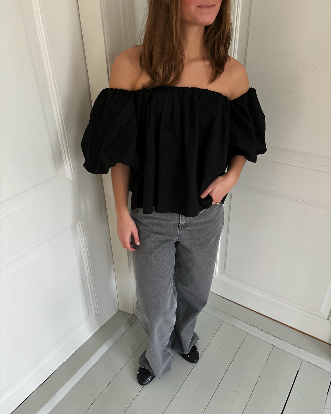 En kvinde står ved en lys væg iført en sort off-shoulder bluse med pufærmer, grå jeans og sorte sko. Hun har hånden i lommen og rører ikke ved væggen.