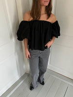 En kvinde står ved en lys væg iført en sort off-shoulder bluse med pufærmer, grå jeans og sorte sko. Hun har hånden i lommen og rører ikke ved væggen.