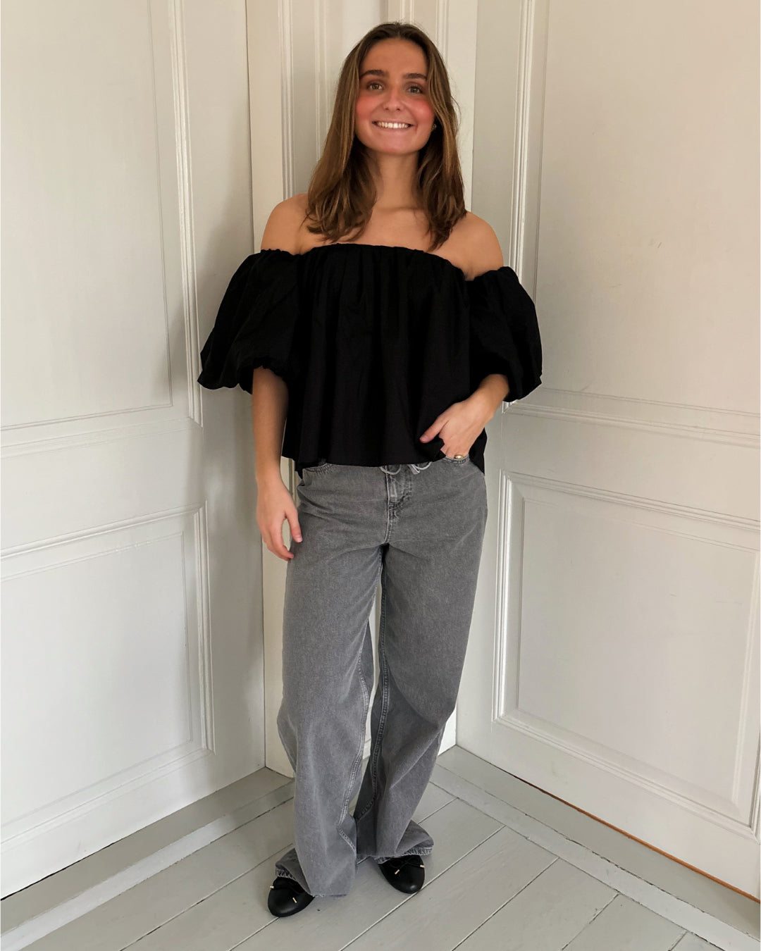 En person står afslappet iført en sort off-shoulder bluse og grå denim bukser i et rum med lyse vægge og gulve.