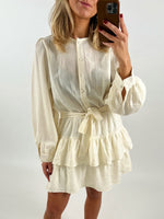 Kvinde tager spejlfoto iført en cremefarvet knap-detaljeret kjole med bæltet talje og flæsekantede lag i et neutralt, lyst rum.