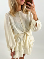 En person iført en lys kjole med knapper og bånd talje, tager selfie. Kjolen har lange ærmer og lagdelte nederdele. Personen står i et neutralt rum.