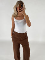 En person tager et spejlfoto med en smartphone. Personen bærer en hvid top og brune bukser i et lyst rum.