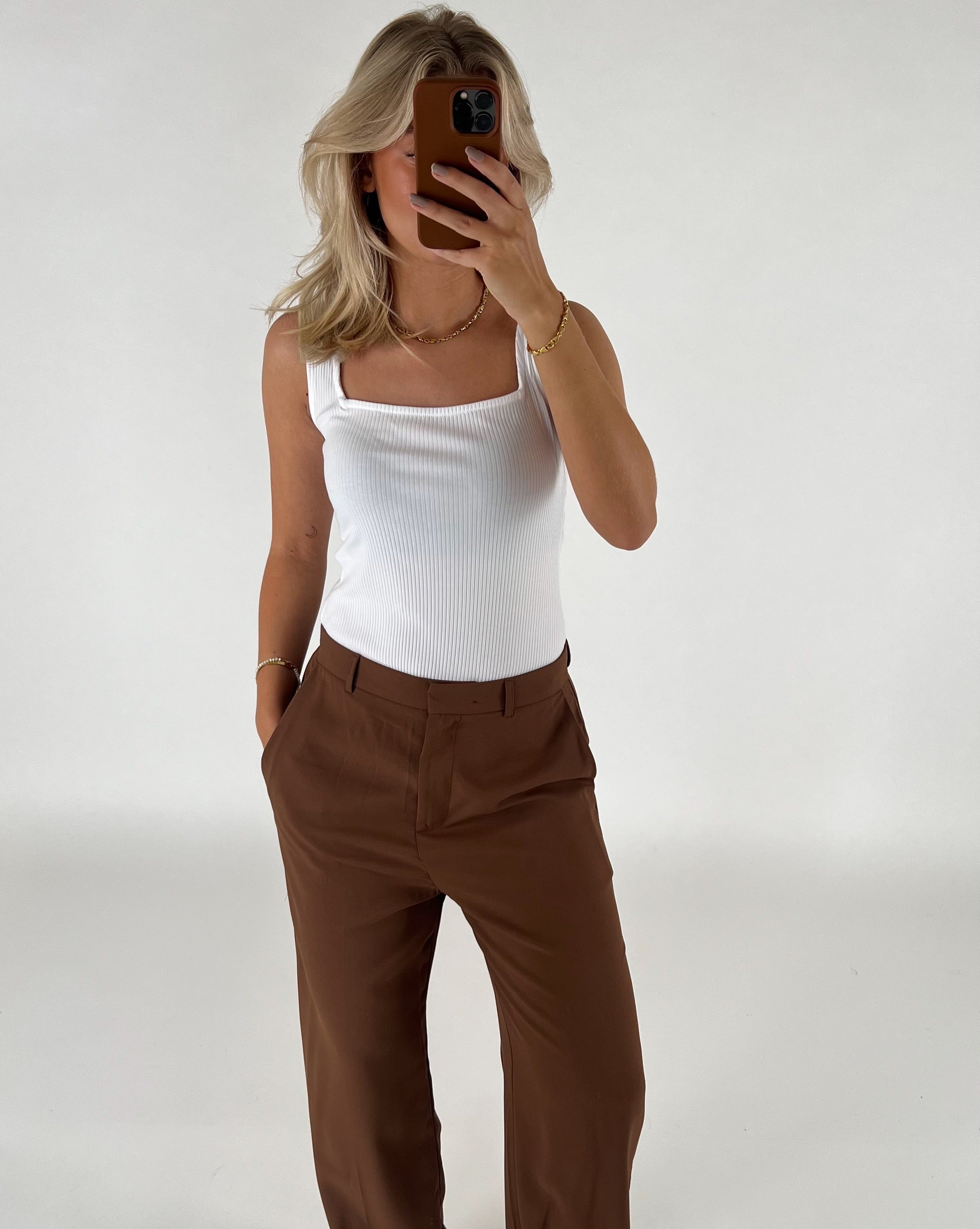 En person tager et spejlfoto med en smartphone. Personen bærer en hvid top og brune bukser i et lyst rum.