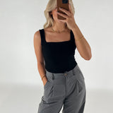 Kvinde i sort top og grå bukser tager et spejlbillede med en smartphone, i en minimalistisk indendørs indstilling med en lys baggrund.