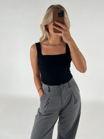 Kvinde i sort top og grå bukser tager et spejlbillede med en smartphone, i en minimalistisk indendørs indstilling med en lys baggrund.