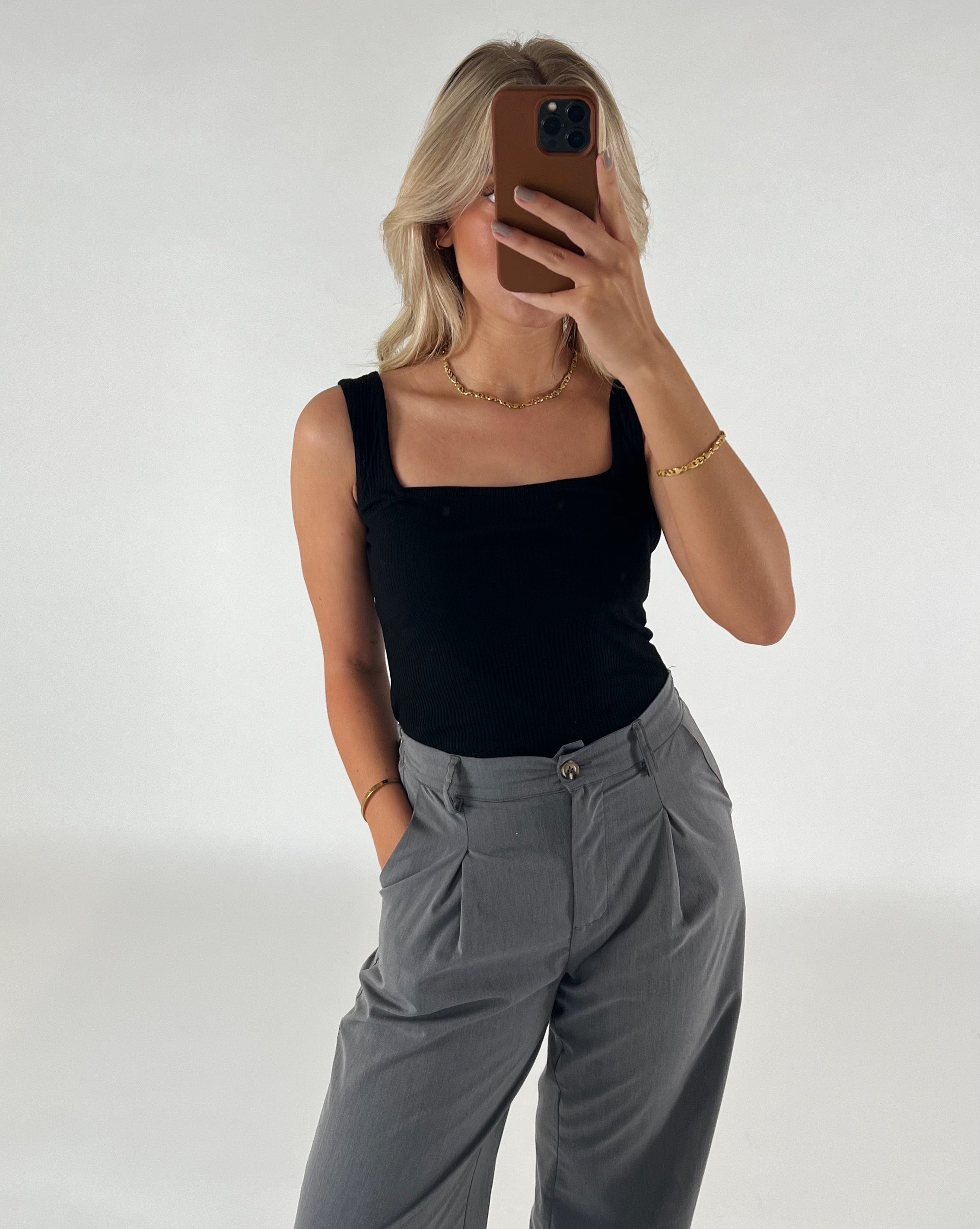 Kvinde i sort top og grå bukser tager et spejlbillede med en smartphone, i en minimalistisk indendørs indstilling med en lys baggrund.