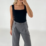 En kvinde i sort top og grå bukser tager et spejlbillede. Hun er omgivet af en neutral, hvid baggrund og bærer guldsmykker.