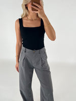 En kvinde i sort top og grå bukser tager et spejlbillede. Hun er omgivet af en neutral, hvid baggrund og bærer guldsmykker.