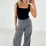 Kvinde tager selfie, iført sort top og grå bukser, står foran en hvid baggrund. Hun har blondt hår og bærer guldsmykker.