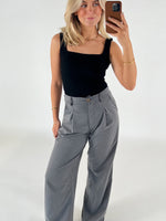 Kvinde tager selfie, iført sort top og grå bukser, står foran en hvid baggrund. Hun har blondt hår og bærer guldsmykker.