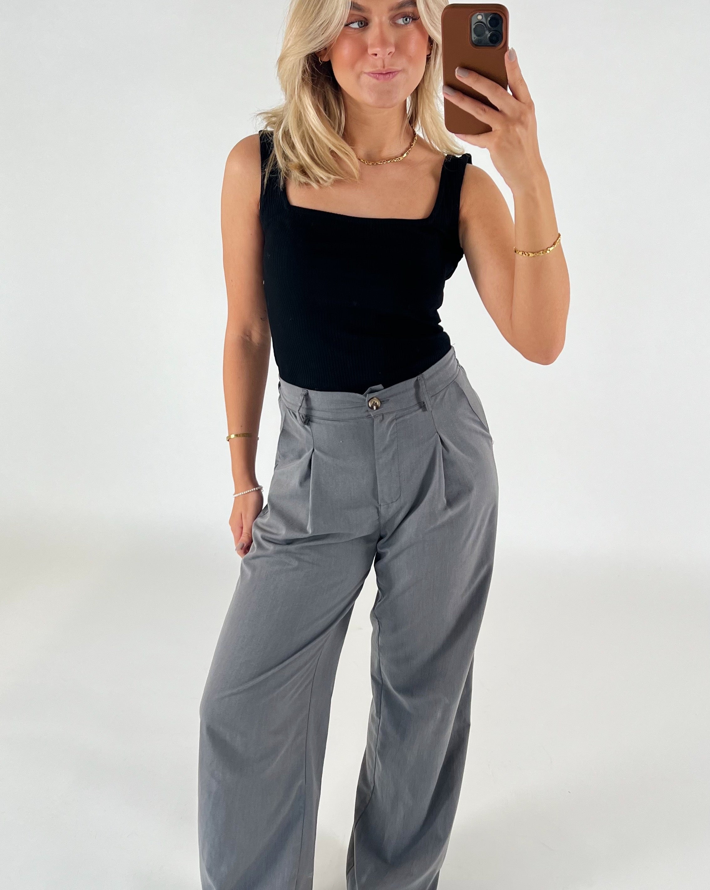 Kvinde tager selfie, iført sort top og grå bukser, står foran en hvid baggrund. Hun har blondt hår og bærer guldsmykker.