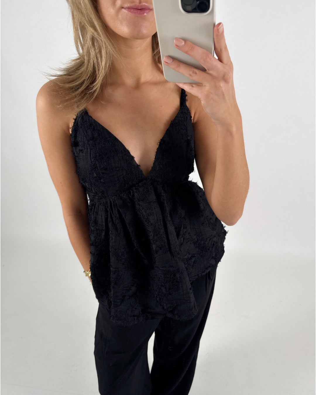 En person iført en sort top med stropper tager et spejlfoto. Personen har lyst hår og står i et lyst rum.