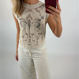 En person i en lysebrun, mønstret striktop og hvide bukser tager et spejlbillede med en smartphone, i et enkelt, lyst rum.