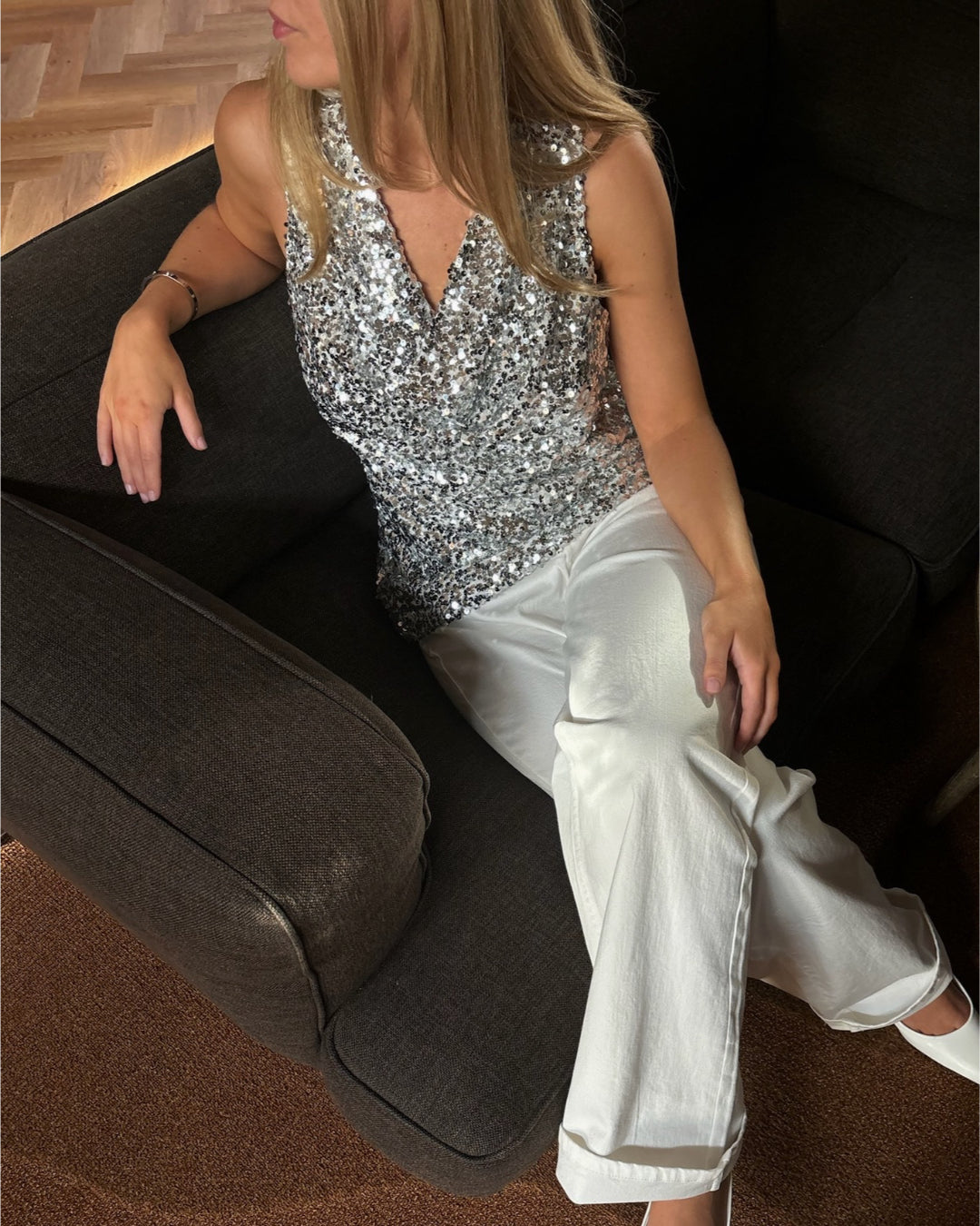 En kvinde iført en sølvglitrende top sidder i en sort lænestol. Hun bærer hvide bukser og har hånden hvilende på armlænet i et rum med trægulv.