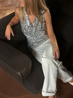 En kvinde iført en sølvglitrende top sidder i en sort lænestol. Hun bærer hvide bukser og har hånden hvilende på armlænet i et rum med trægulv.