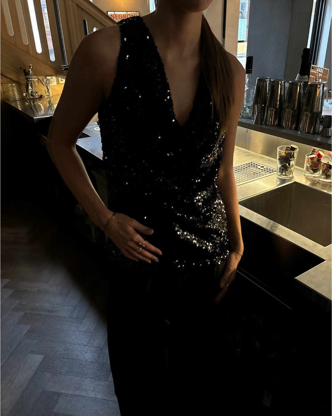 En person iført en sort, glitrende top står ved en bar med metaloverflader og cocktailshakere i baggrunden. Skiltet "Negroni" ses svagt på væggen.