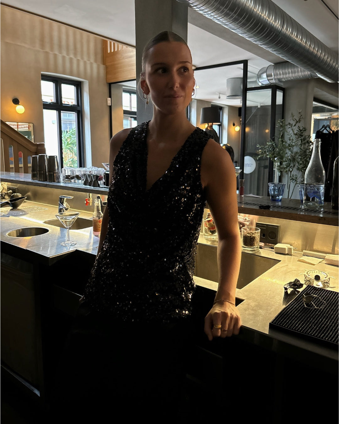 En person iført en glitrende sort top står ved en bar, omringet af glas og flasker, i en stilfuld café med dæmpet belysning og moderne indretning.