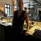 En person iført en glitrende sort top står ved en bar, omringet af glas og flasker, i en stilfuld café med dæmpet belysning og moderne indretning.