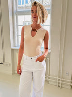 En kvinde iført en lyseblå top og hvide bukser står afslappet i et lyst rum med store vinduer og synlige rør langs væggen.