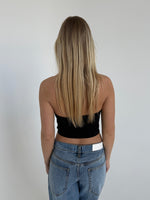 Kvinden har langt, lyst hår og bærer en sort, stropløs top samt blå jeans. Hun står med ryggen til i et rum med en ensfarvet, hvid væg.