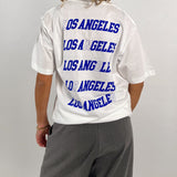 En person bærer en hvid T-shirt, hvorpå der står "LOS ANGELES" gentaget fire gange i blå skrift. Personen står med ryggen til i et neutralt rum.