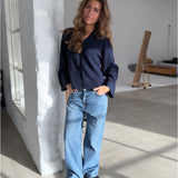 Kvinden står afslappet op ad en hvid mur, iført en mørk trøje og lyse jeans. Rummet har store vinduer og moderne indretning med minimalisme som stil.