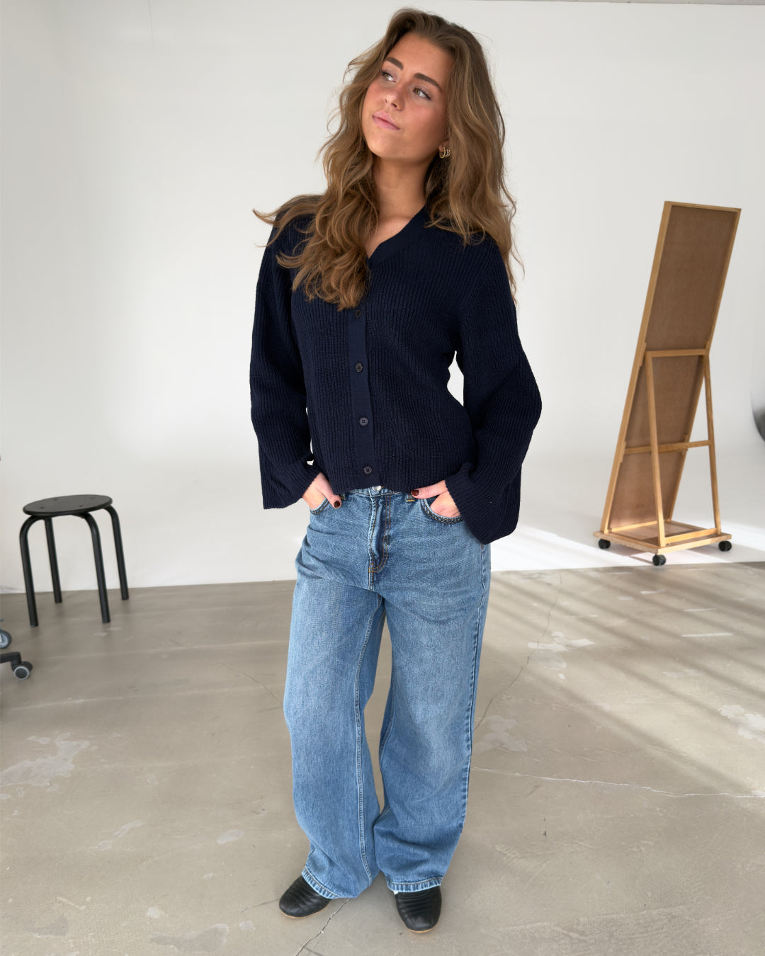 En person står afslappet med hænderne i lommerne iført en mørk striktrøje og blå jeans. Baggrunden inkluderer en skammel og et staffeli i et lysegråt rum.