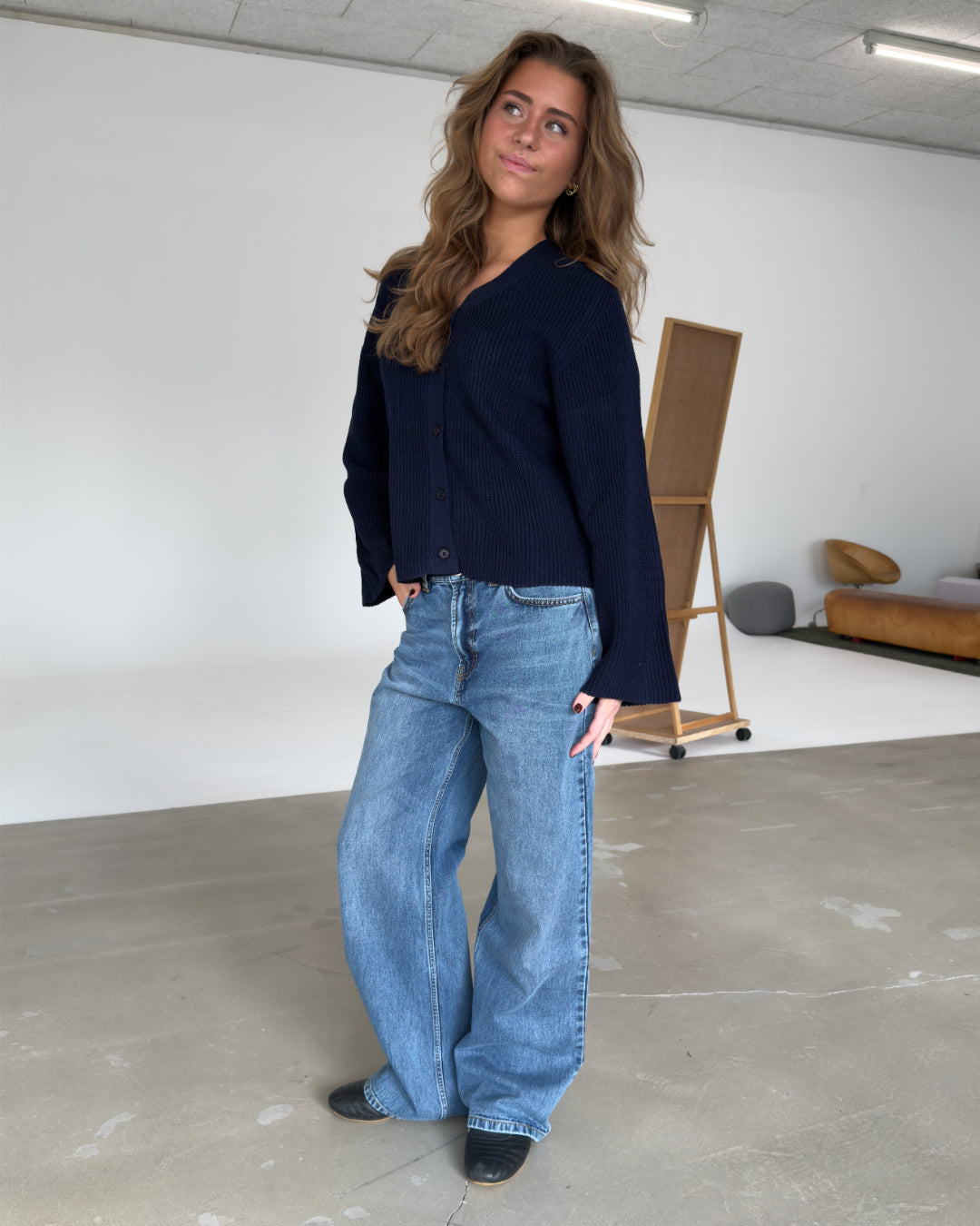 En person i en mørk bluse og brede jeans står afslappet i et rummeligt, minimalistisk rum med en lysebrun sofa og en staffeli i baggrunden.