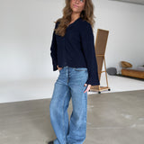 En person i en mørk bluse og brede jeans står afslappet i et rummeligt, minimalistisk rum med en lysebrun sofa og en staffeli i baggrunden.