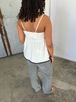 En person med krøllet hår står med ryggen til, iført en hvid top og grå bukser, på et betonlignende gulv foran en rusten metalvæg.