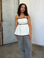 En person iført en hvid top med sorte kanter står afslappet foran en metaldør i et rum med beton-gulv, ser til siden med et eftertænksomt udtryk.