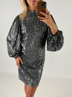 En person iført en glitrende sølvkjole tager en selfie med en smartphone. Personen står i et neutralt, indendørs miljø.