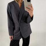 En person iført en mørk blazer tager et spejlbillede med en smartphone. Personen står i et minimalistisk, lyst rum uden synlige dekorationer.