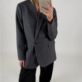 En person iført en mørk blazer tager et selfie med en smartphone. Personen står i et minimalistisk, hvidt rum.