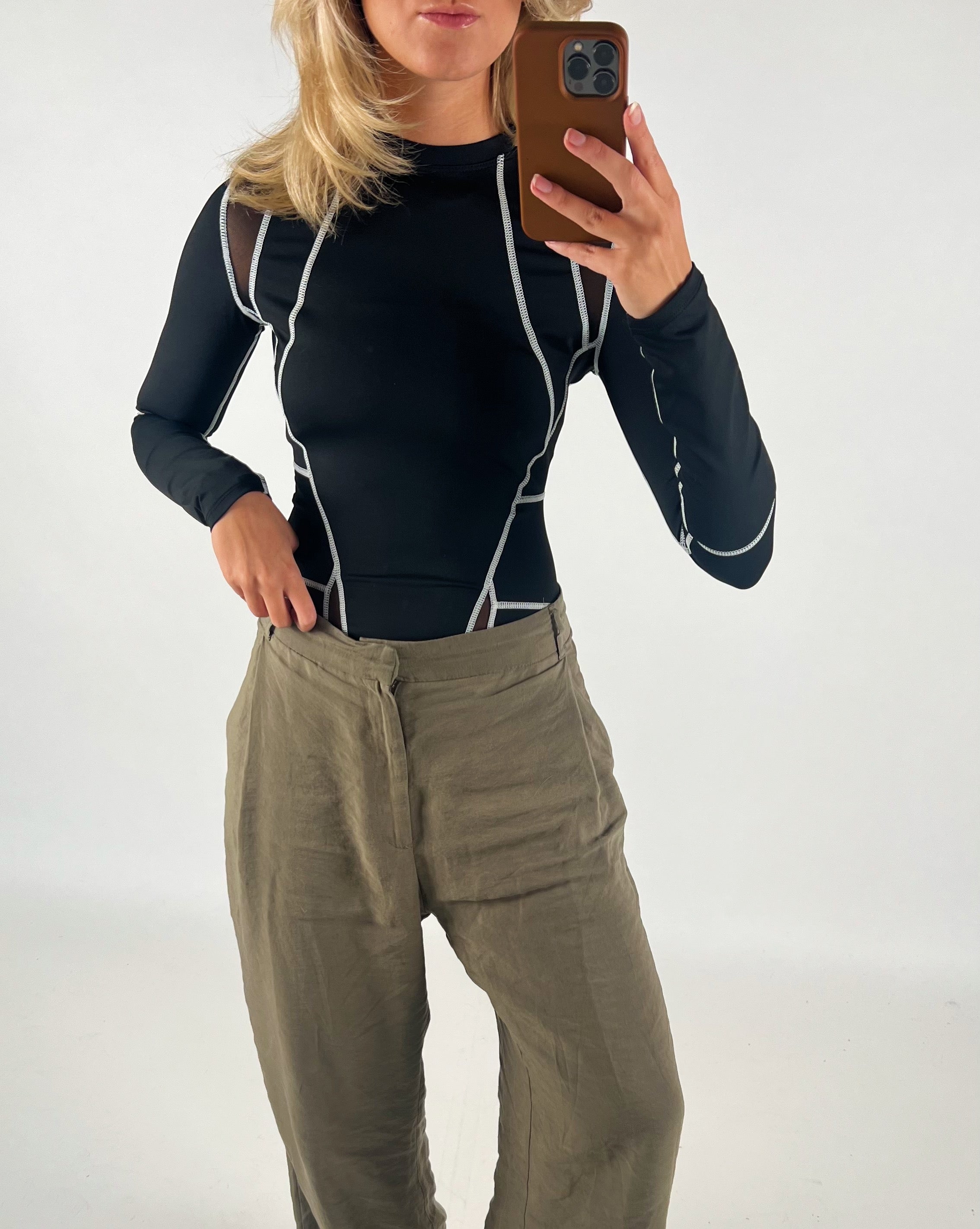 En person i sort trøje med hvide sømme tager en selfie, mens de står i beige bukser mod en hvid baggrund.