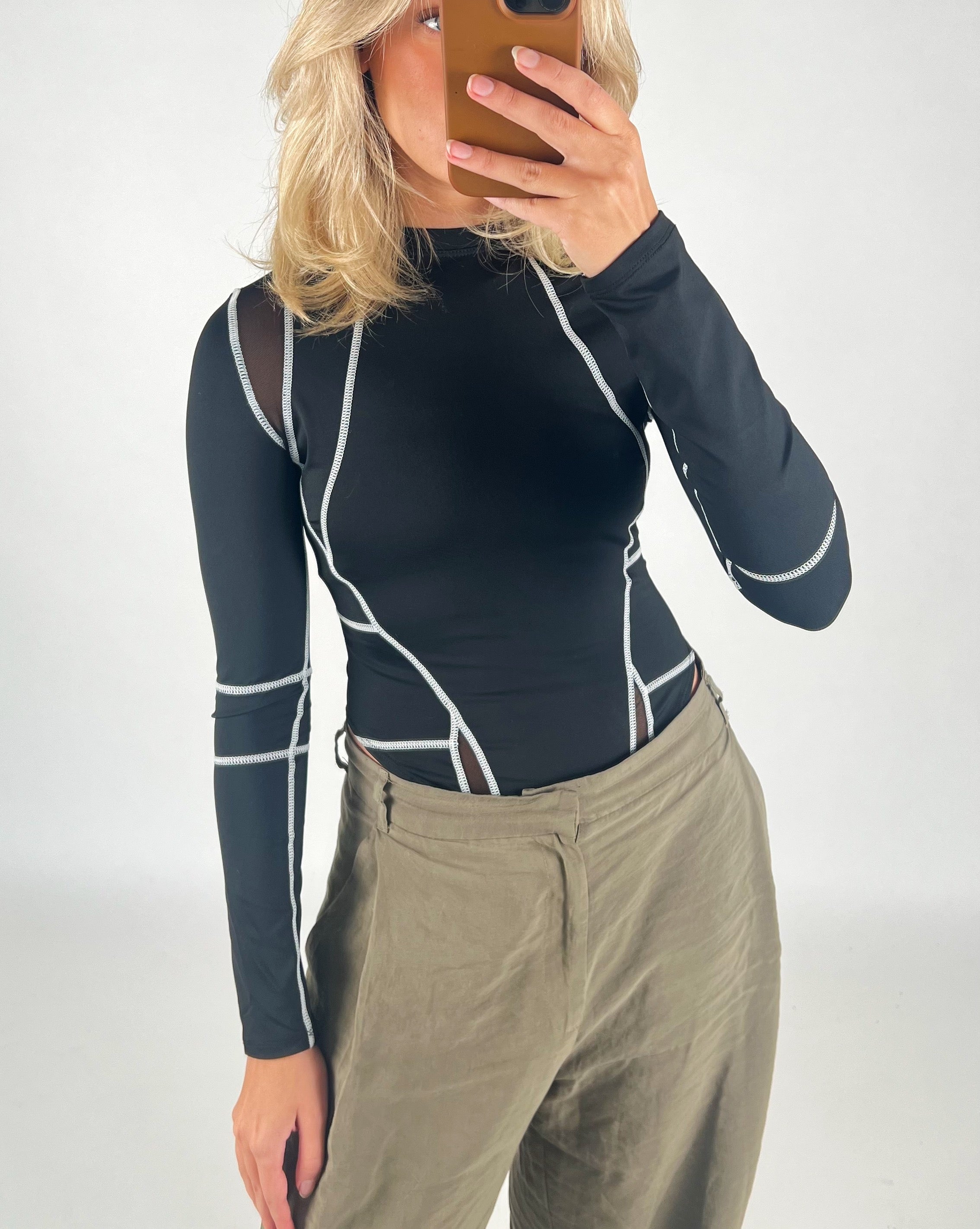 En person iført en sort trøje med hvide sømme tager en selfie i et spejl. Personen har lyse bølgende hår og bærer olivengrønne bukser i en lys omgivelser.