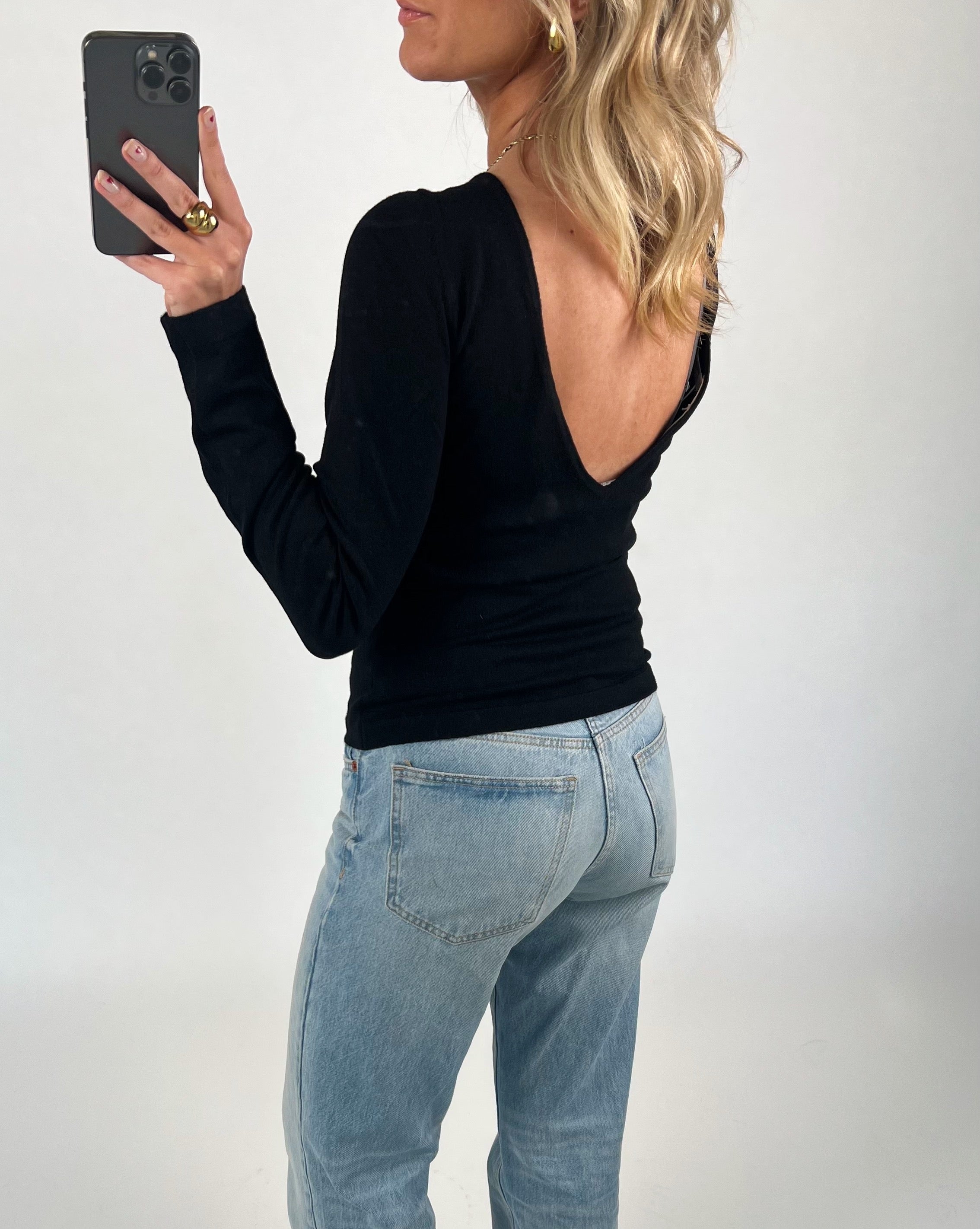 En person iført en sort bluse med åben ryg tager et spejlbillede med en smartphone. De bærer lyseblå jeans i et neutralt oplyst rum.