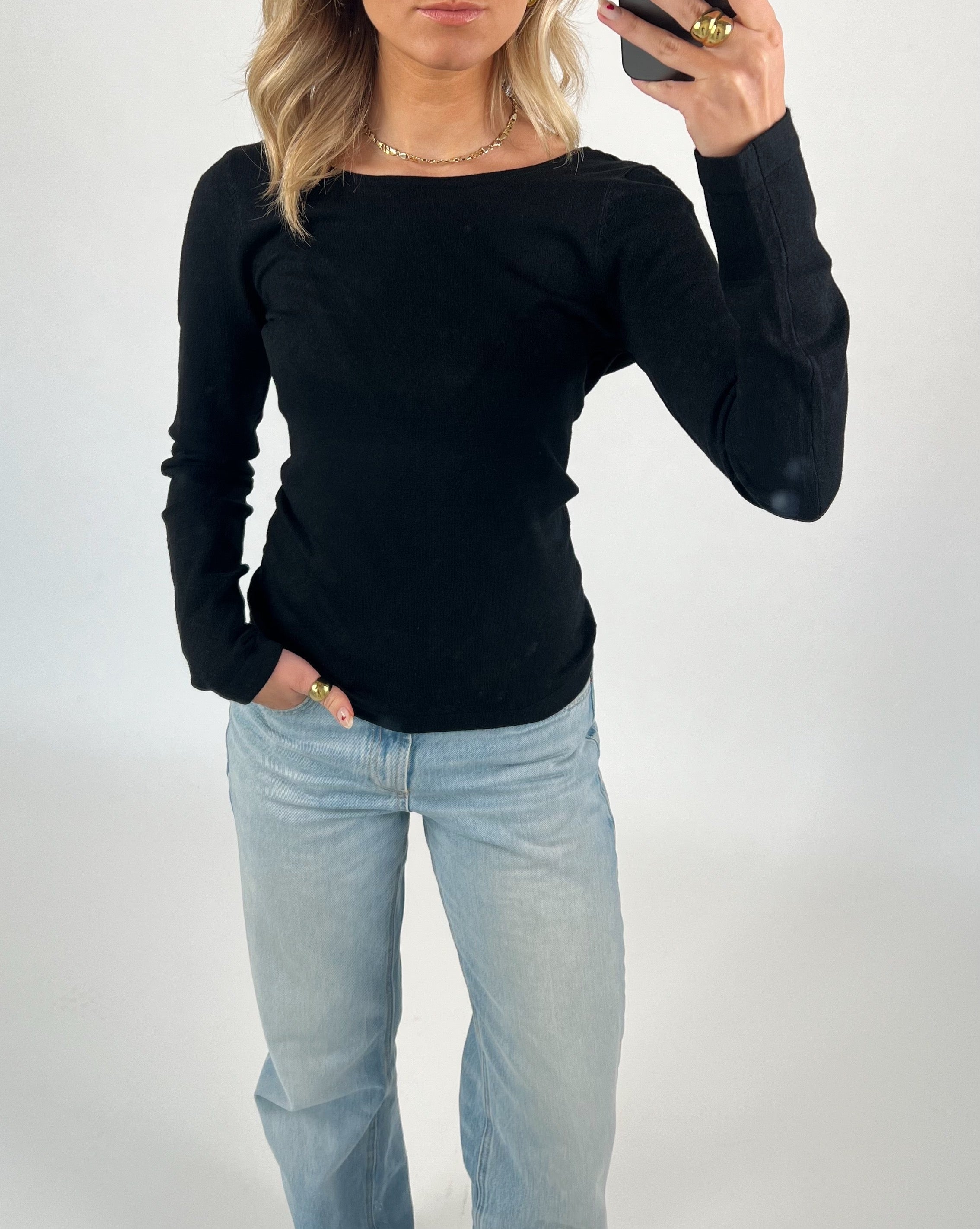 Kvinde iført sort trøje og lyseblå jeans, tager selfie med telefon. Baggrund er hvid og enkel. Hendes hånd med ring og neglelak ses i forgrunden.
