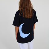 En person iført en sort T-shirt med en blå måne på ryggen står med ryggen til og har langt, brunt hår. De har hvide bukser på i et neutralt rum.
