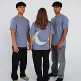 Tre personer står i en lys baggrund. To mænd i grå T-shirts vender mod kameraet, og en kvinde i lignende T-shirt med en hvid måne på ryggen står med ryggen til.