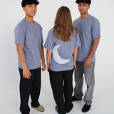 Tre personer står sammen i en cirkel, iført grå t-shirts. En person vender ryggen til, med en måneform på trøjen. De er i et minimalistisk, hvidt rum.