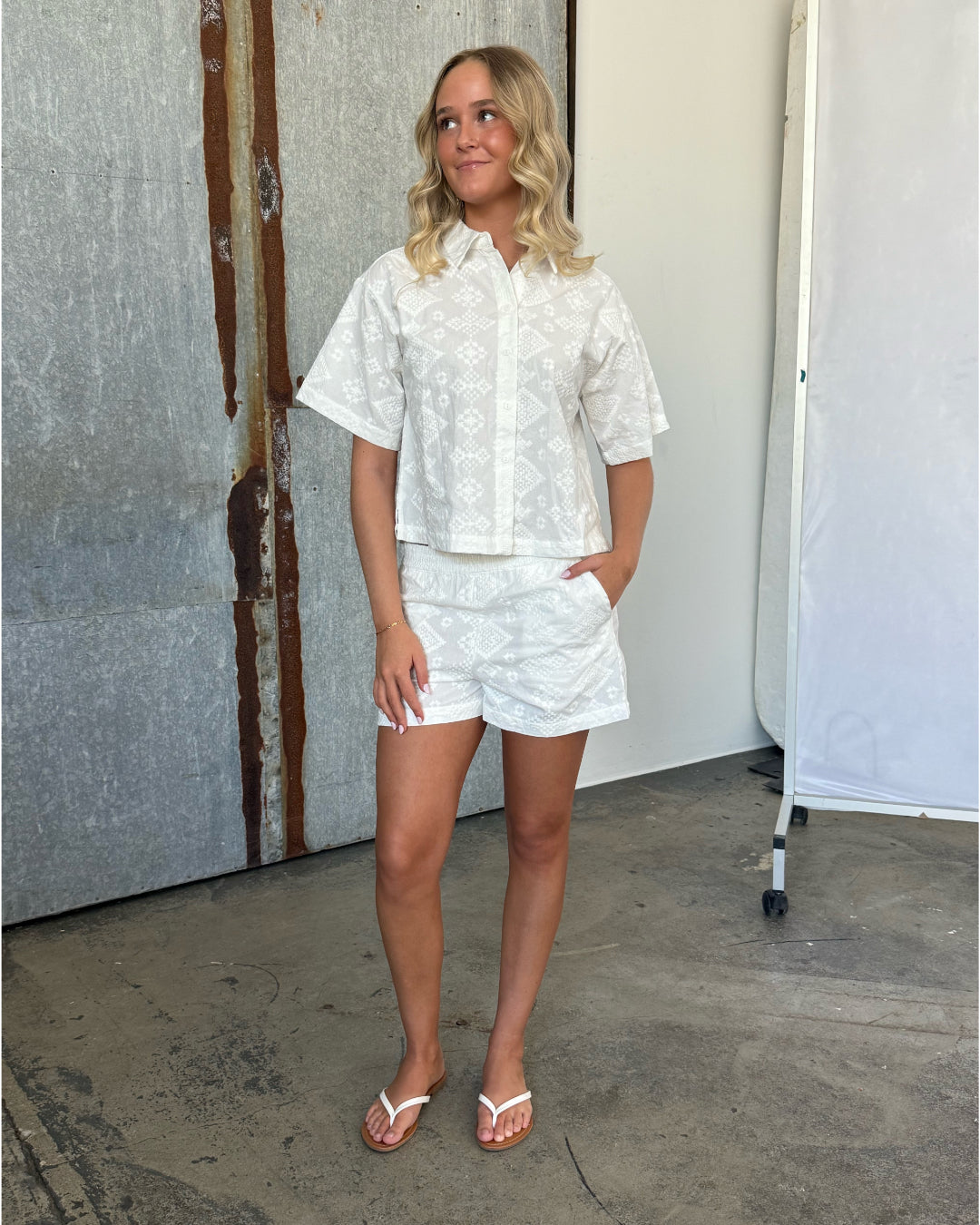 En person iført hvidt mønstret tøj og sandaler står afslappet med hånden i lommen. Baggrunden består af en rustik metalvæg og en lys skillevæg.